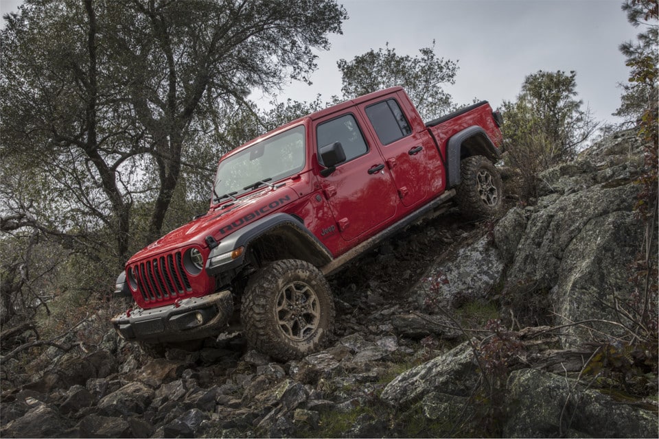 Camion Jeep Rubicon rouge roulant sur un terrain rocheux, entouré d'arbres et de paysages accidentés.