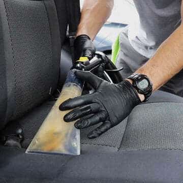 Personne utilisant un aspirateur pour nettoyer un siège de voiture, portant des gants noirs et une montre, mettant en valeur l'entretien de l'intérieur d'une voiture.