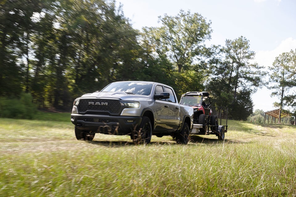 RAM truck towing an ATV on a trailer through a grassy field, surrounded by trees.