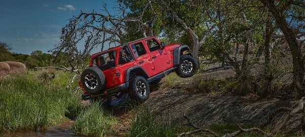 A red off-road vehicle navigating rugged terrain with greenery and trees under a clear sky.