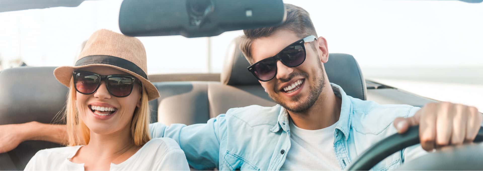 Un couple souriant avec des lunettes de soleil conduisant dans une décapotable par une journée ensoleillée.
