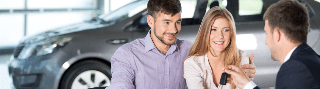 Un couple souriant discute des options d'achat d'une voiture avec un vendeur, à côté d'un véhicule dans un concessionnaire.