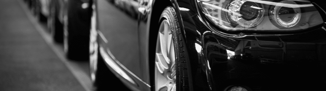Gros plan de la roue avant et du phare d'une voiture garée, reflétant les lumières de la rue urbaine, en noir et blanc.