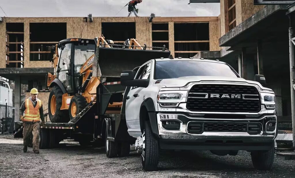 A white RAM truck towing construction equipment at a building site with workers nearby.
