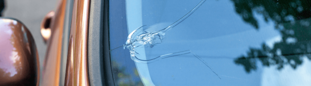 Pare-brise de voiture fissuré avec des dommages d'impact visibles près du rétroviseur.