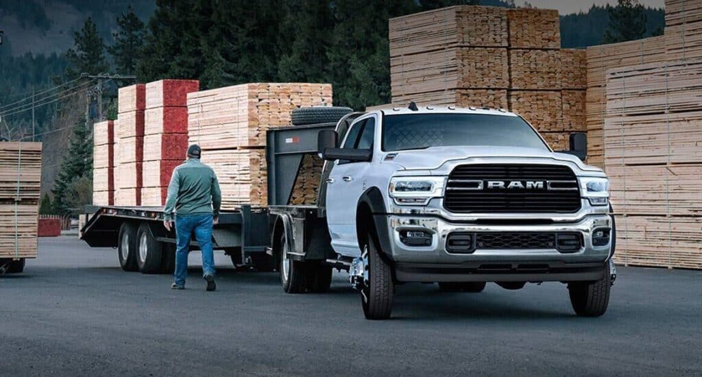 A white RAM truck towing a trailer loaded with lumber, parked in a timber yard.