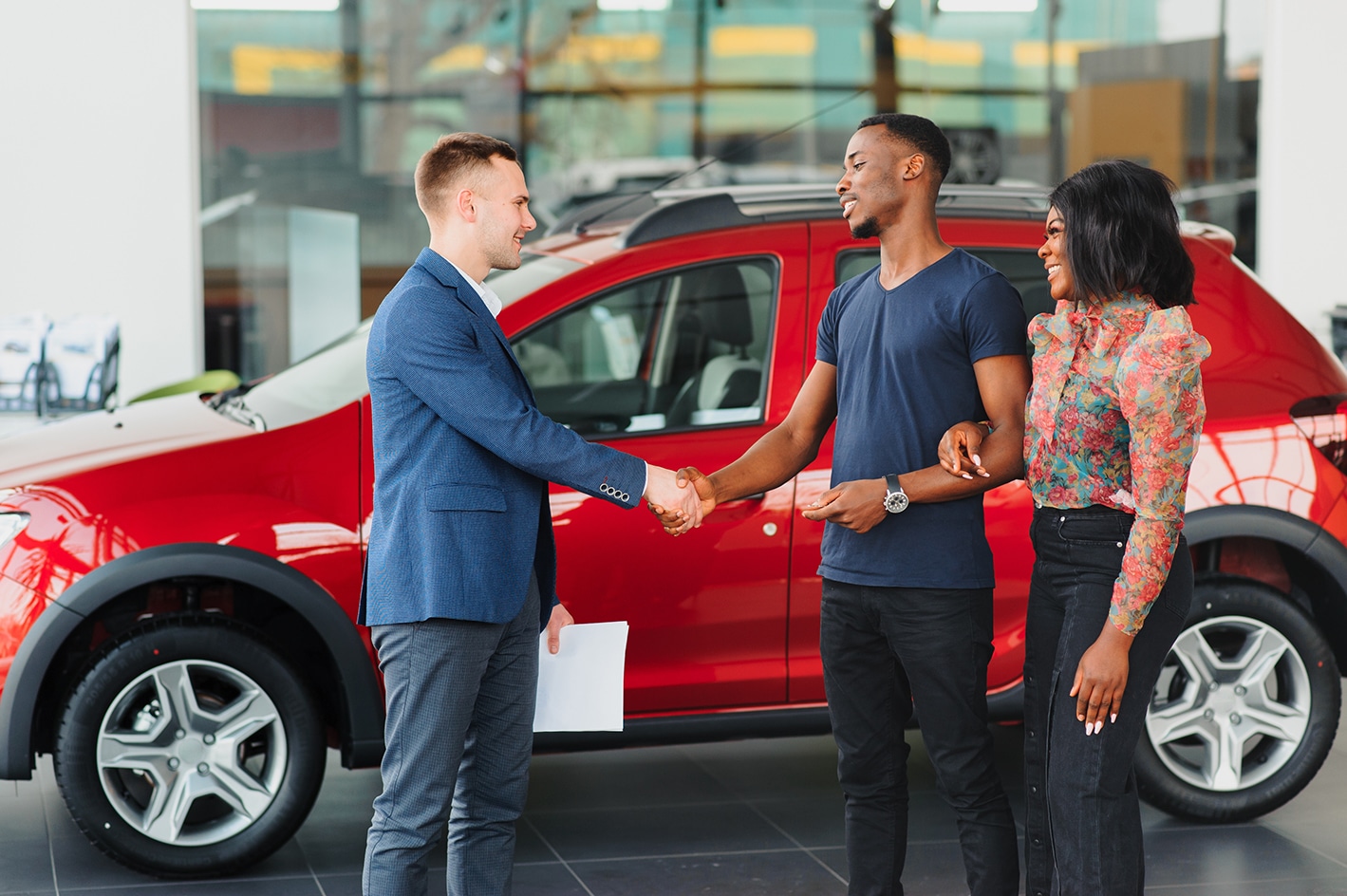 Vendeur de voiture serrant la main d'un couple souriant devant un VUS rouge dans une concession.