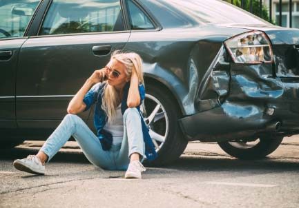 Woman sitting by damaged car, talking on phone after accident, appearing distressed.