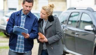 A man shows a clipboard to a woman near a parked car, likely discussing car details or paperwork.
