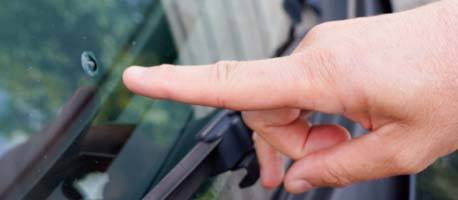 A finger pointing at a small chip on a car windshield, indicating damage.