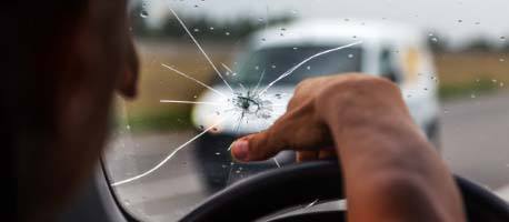 Cracked windshield with driver's hand on steering wheel, car approaching on road.