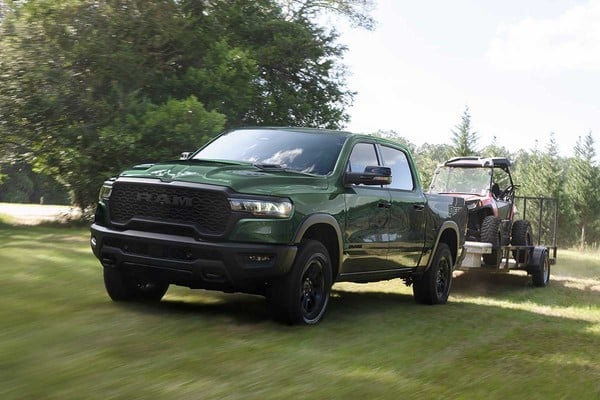 Green pickup truck towing an off-road vehicle on a trailer, driving through a grassy area with trees.