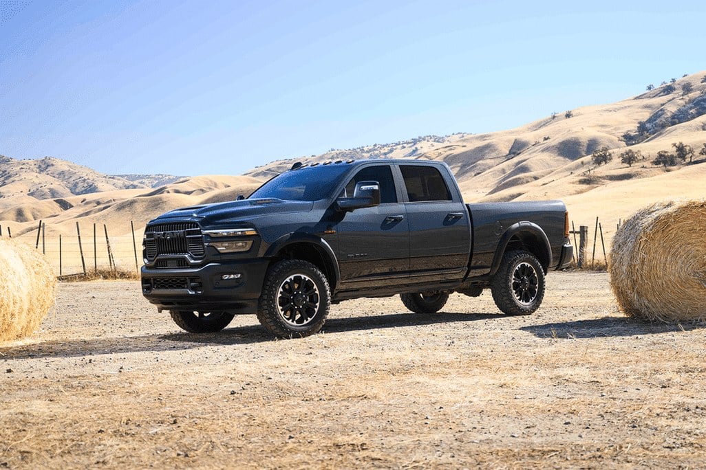 Black Ram 2500 pickup truck parked on a gravel ranch lot between hay bales, golden hills behind.